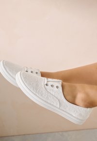 Baskets en dentelle blanche avec un motif floral, bout rond, œillets métalliques, tige texturée et semelle en caoutchouc blanche.