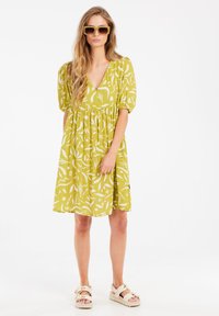 Grünes blumengemustertes Kleid mit kurzen Puffärmeln und V-Ausschnitt. Texturiertes Material und lockere Passform, kombiniert mit hellen Sandalen.
