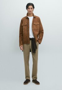 Braune Wildlederjacke mit zwei vorderen Taschen, darunter ein weißes T-Shirt, beige Hose und dunkelbraune Schuhe. Das Model steht vor einem hellen Hintergrund.