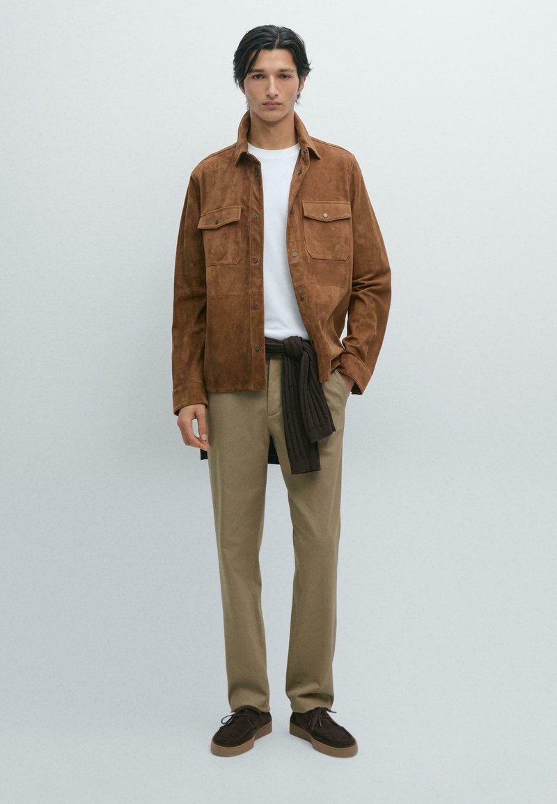 Braune Wildlederjacke mit zwei vorderen Taschen, darunter ein weißes T-Shirt, beige Hose und dunkelbraune Schuhe. Das Model steht vor einem hellen Hintergrund.