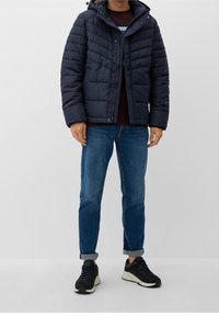 Laivastonsininen toppatakki, jossa vaakasuora tikattu kuvio, vetoketjusuljenta ja huppu. Yhdistetty sinisiin farkkuihin ja mustiin lenkkareihin, neutraalilla taustalla.