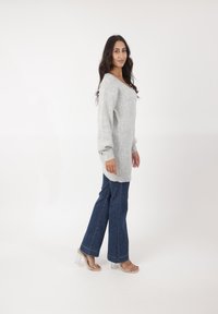 Pull en tricot gris avec un col en V, manches longues, associé à un jean bleu évasé et des sandales à talons blocs transparentes. Fond simple.