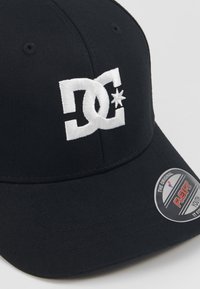 Gorra de béisbol negra de tela con un logotipo bordado blanco y un diseño de estrella. La etiqueta cosida indica tamaño "Flexfit Youth".