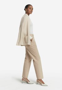 Beige kofta, vit åtsittande topp, ljusbeiga byxor och spetsiga slingback-klackar med en texturmönster och svarta accenter.