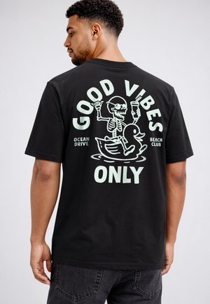 Mann trägt schwarzes T-Shirt mit Skelett, das ein Getränk hält und auf einer aufblasbaren Ente reitet, Text "Good Vibes Only, Ocean Drive Beach Club" auf dem Rücken.