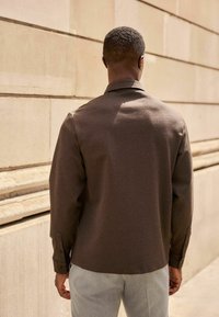 Căma�șă maro cu guler, cu o textură netedă și manșete cu nasturi, combinată cu pantaloni gri deschis, privită din spate, pe un fundal neutru.