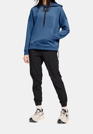 Osoba ubrana w niebieską bluzę z kapturem, czarne spodnie dresowe, białe skarpetki i białe buty do biegania na neutralnym tle.