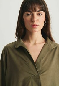 Chemise verte olive à col, avec un design en V, fabriquée dans un tissu lisse. Présente une coupe décontractée et des détails de couture subtils au niveau de la poitrine.
