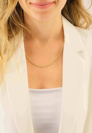 Collana di catena dorata indossata sopra un top bianco, con un design attorcigliato. Il modello ha capelli ondulati e chiari ed è vestito con un blazer bianco.