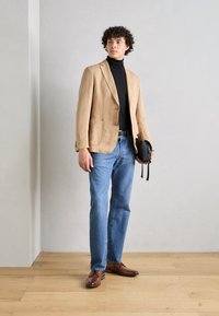 Lichtbruin linnen blazer, zwarte coltrui, blauwe jeans en bruine leren schoenen. Houdt een kleine zwarte tas vast, staande op een houten vloer.