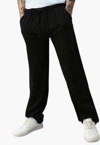 Schwarze Jogginghose mit elastischem Bund und Kordelzug, ausgestattet mit Seitentaschen. Kombiniert mit weißen Turnschuhen auf einem schlichten Hintergrund.