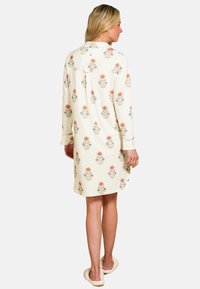 Vrouw in een beige jurk tot de knie met lange mouwen en een bloemenpatroon, staat blootvoets tegen een witte achtergrond.