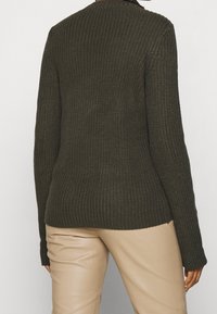 Pull vert foncé en tricot avec un motif texturé, manches longues et encolure arrondie, associé à un pantalon beige.