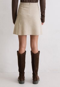 Person trägt einen beigen Rock und dunkelbraune kniehohe Wildlederstiefel und steht vor einem einfarbigen weißen Wandhintergrund.