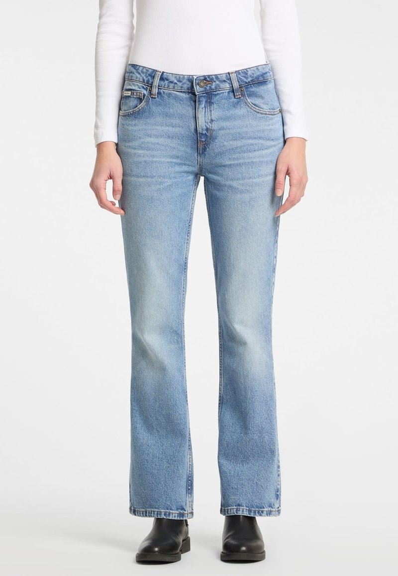 Personne portant un jean bootcut bleu clair, une chemise blanche à manches longues et des bottines noires, debout devant un fond uni.