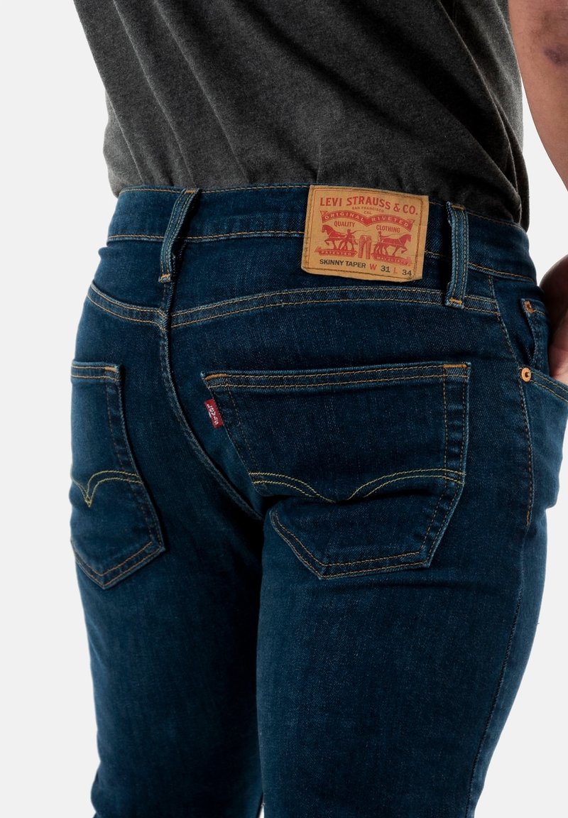 Dunkelblaue Skinny-Jeans mit gelben Nähten, ausgestattet mit einem Lederpatch mit Branding an der Taille und traditionellem Rücktaschendesign.
