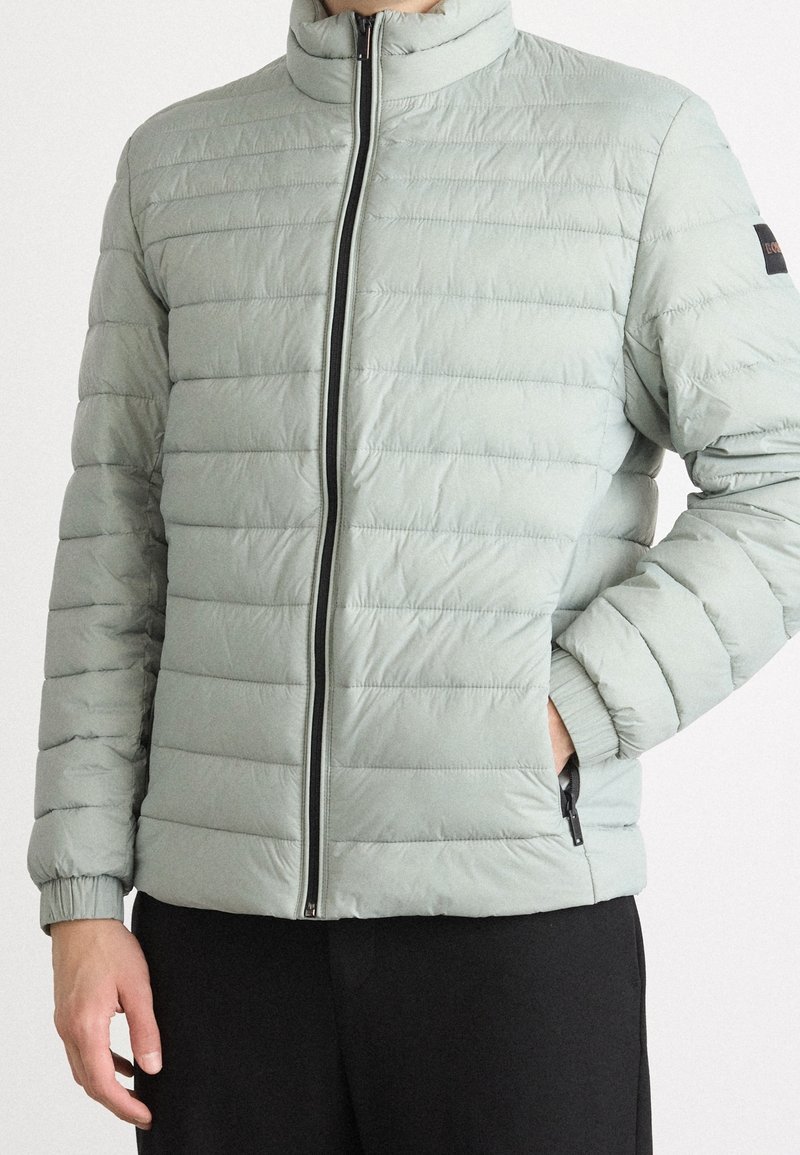 Homme portant une veste matelassée gris clair avec une fermeture éclair noire et une main dans la poche, associée à un pantalon noir.
