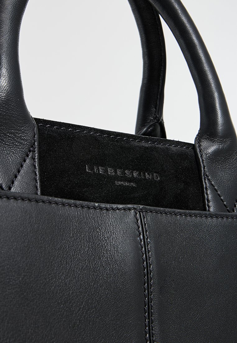 Schwarze Lederhandtasche mit glatter Textur, kontrastierender Wildleder-Panel und geprägtem Logo auf einer rechteckigen Basis. Kurze, runde Griffe für einen sicheren Halt.