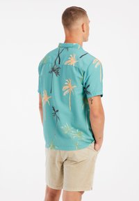 Kortekleurige blouse in turquoise stof met palmboomprint in geel en zwart. Bevat een button-down kraag en een relaxte pasvorm.