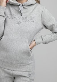 Mujer vistiendo un hoodie gris claro de SikSilk y pantalones de chándal a juego, con la mano derecha en el bolsillo y la mano izquierda cerca de la capucha.