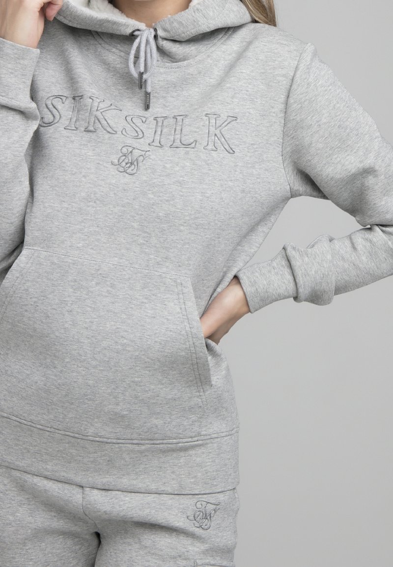 Mujer vistiendo un hoodie gris claro de SikSilk y pantalones de chándal a juego, con la mano derecha en el bolsillo y la mano izquierda cerca de la capucha.