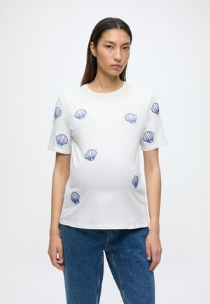 Kvinde med langt mørkt hår iført en hvid T-shirt med blå muslingetryk og blå jeans, stående mod en ensfarvet hvid baggrund.