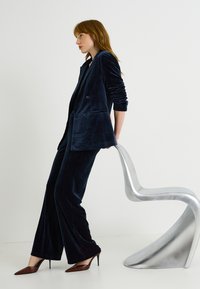 Traje de terciopelo azul marino con blazer cruzado y pantalones acampanados. La modelo se apoya en una elegante silla metálica. Tacones marrones con punta afilada.