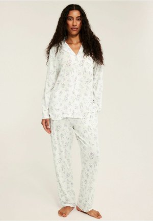 Vrouw staat op blote voeten met een wit, bloemenprint, pyjamaset met lange mouwen, een knoopsluitend bovenstuk en loszittende broek tegen een effen achtergrond.