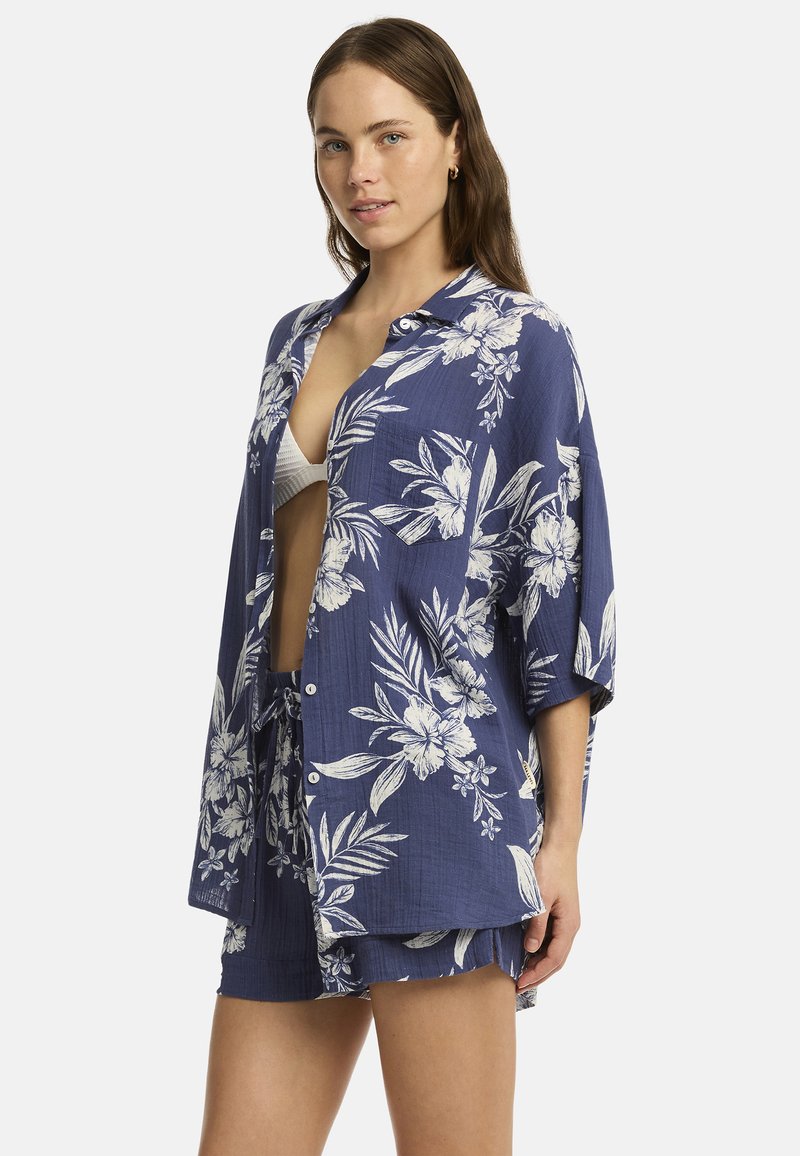 El modelo posa con una camisa de botones navy con estampado floral y pantalones cortos a juego, mostrando un estilo relajado con un bikini blanco debajo.