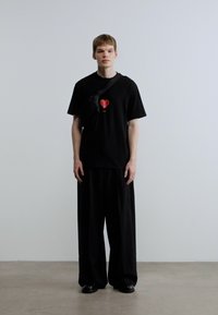 Μαύρο κοντομάνικο μπλουζάκι με γραφικό κόκκινης καρδιάς, σε συνδυασμό με φαρδιά μαύρη παντελόνια και μαύρη τσάντα χιαστί. Το σύνολο ολοκληρώνουν τα επίπεδα παπούτσια.