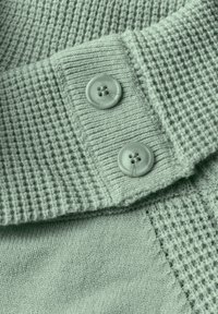 Tyrkysová pletenina se vzorem vafle, s hladkými částmi a dvěma odpovídajícími kulatými knoflíky se čtyřmi dírkami, ve velkém přiblížení.