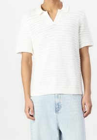 Biała, dziana koszulka polo z krótkim rękawem i dekoltem w serek, z poziomymi fakturowanymi paskami i prążkowanym wykończeniem. Noszona z jasnoniebieskimi dżinsami.