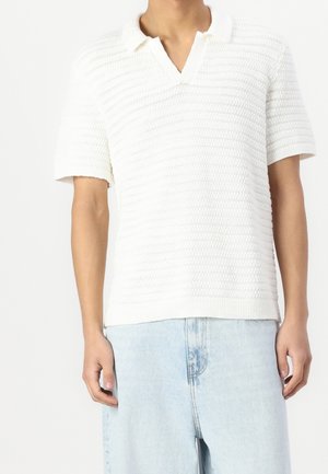 Witte gebreide polo met korte mouwen en V-hals, voorzien van horizontale textuurstrepen en een geribde onderkant. Gemaakt in combinatie met lichtblauwe jeans.