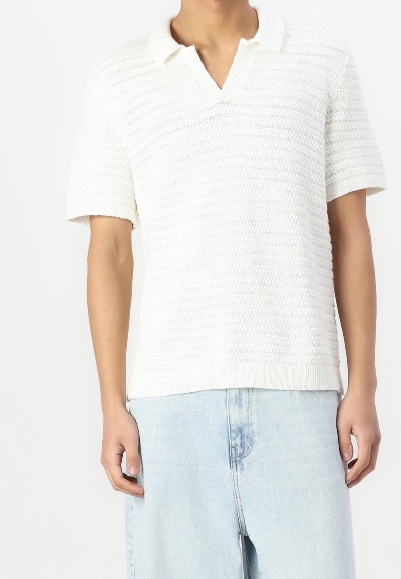 Biała, dziana koszulka polo z krótkim rękawem i dekoltem w serek, z poziomymi fakturowanymi paskami i prążkowanym wykończeniem. Noszona z jasnoniebieskimi dżinsami.