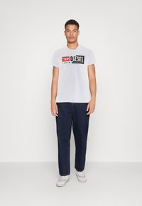  camiseta blanca de manga corta con el logo "DIESEL" en negro y rojo, combinada con pantalones de mezclilla azul oscuro, exhibida sobre un fondo blanco plano.
