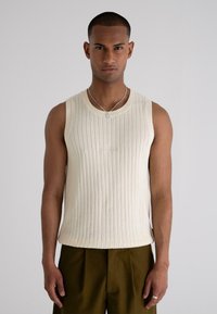 Cream ribbed sleeveless sweater with a round neckline and subtle embossed detail, paired with olive green pants. Smooth texture and fitted design.