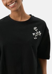 Camiseta negra de manga corta con un diseño floral en rhinestones plateados en el lado izquierdo, con un corte relajado y cuello redondo.