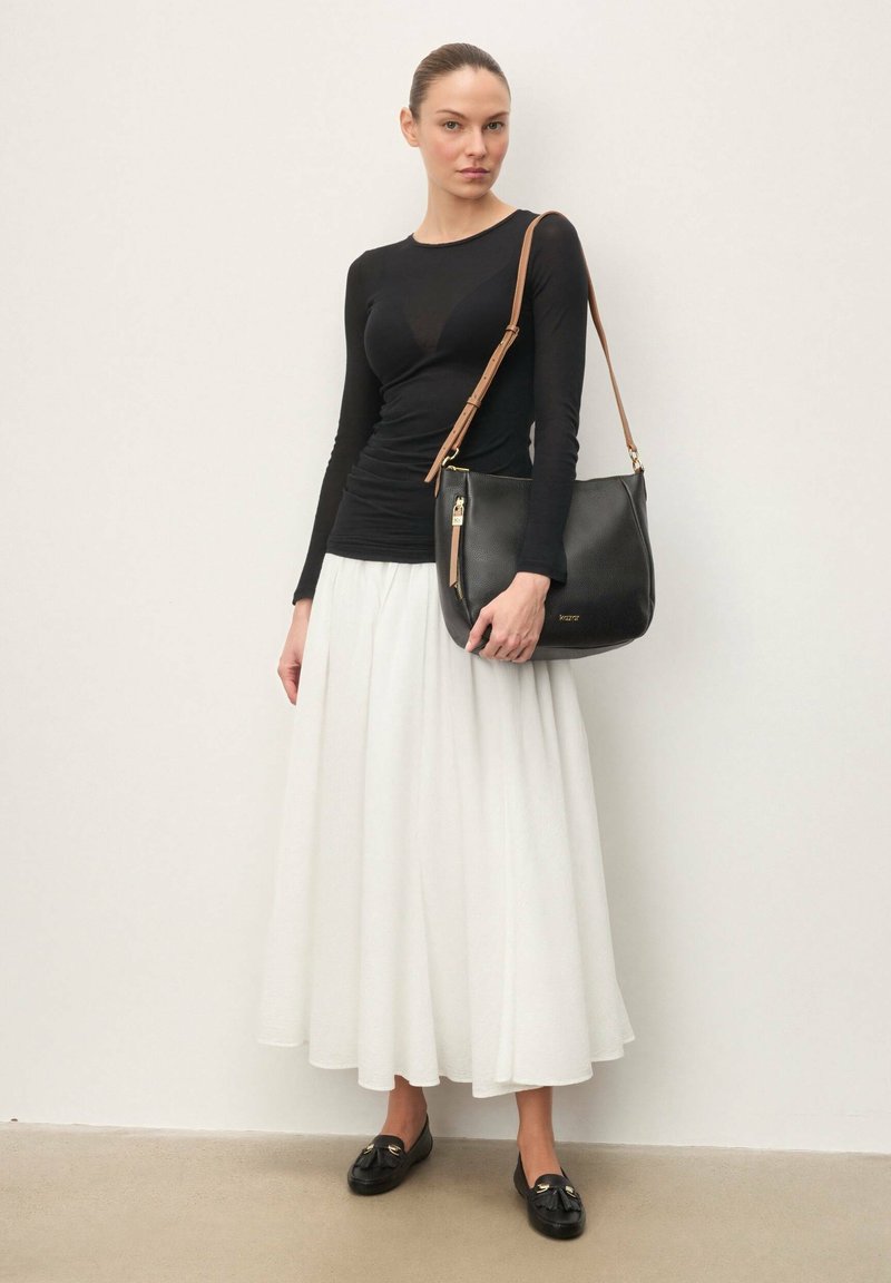 Femme portant un haut à manches longues noir, une jupe midi blanche, des mocassins noirs, tenant un sac à épaule noir avec une sangle beige contre un mur uni.