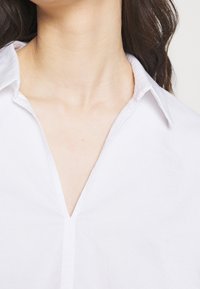 Camicia bianca con colletto a v, caratterizzata da una texture liscia e linee pulite. Il tessuto appare leggero e fresco.