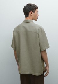 Camicetta a maniche corte color verde oliva realizzata in lino, con una vestibilità comoda, un colletto piccolo e spacchi laterali all'orlo per facilitare il movimento.