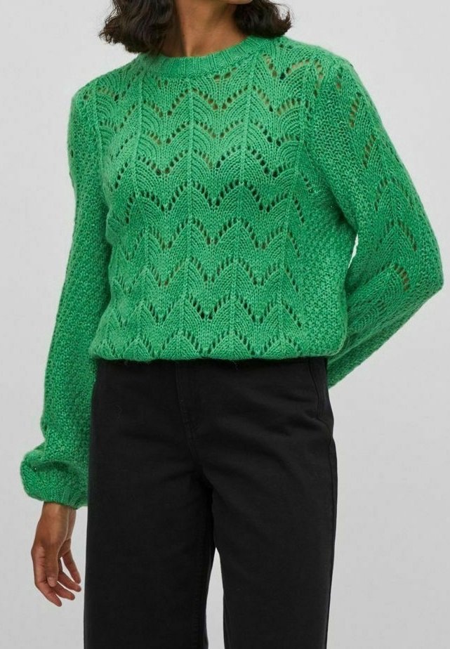 Femme portant un pull en maille vert vif à manches longues avec un motif en dentelle chevron, associé à un pantalon noir taille haute.