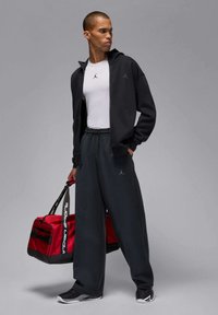 Zwarte hoodie met ritssluiting, wit T-shirt met logo, donkergrijze joggingbroek, een rode sporttas vasthoudend. Gladde stof, comfortabele pasvorm, sportief ontwerp.