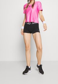 Camiseta deportiva corta de color rosa con logo negro, combinada con pantalones cortos deportivos negros que tienen una cinturilla blanca, y zapatillas negras con detalles blancos.