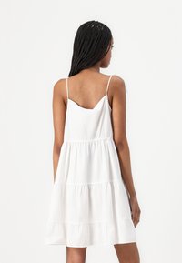 White cotton dress with thin straps, tiered design, and a flowing silhouette. The back features a V-neckline and gathered fabric detail.
