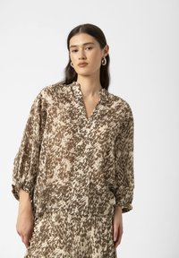 Braun- und cremefarbige Bluse mit Muster, leichtes Material, V-Ausschnitt, lockerer Schnitt und weit ausgestellte Ärmel. Auffälliges abstraktes Design auf der gesamten Fläche.