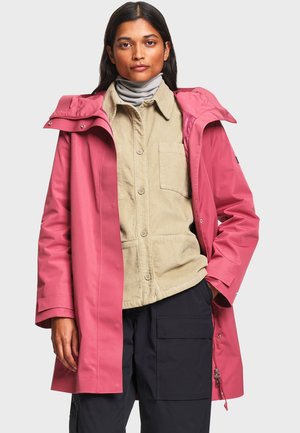 Femme portant un imperméable rose par-dessus une chemise beige boutonnée et un col roulé gris avec un pantalon cargo noir, debout devant un fond uni.