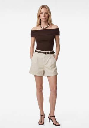 Vrouw draagt een bruin off-shoulder top, beige short met riem, parelketting en zwarte sandalen met hak, staande met handen in de zakken.