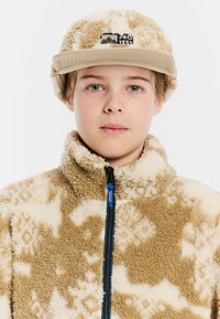 Chaqueta de forro polar en beige con patrones crema; cuello alto y cremallera azul. Gorro beige a juego con logo. Apariencia texturizada y suave.