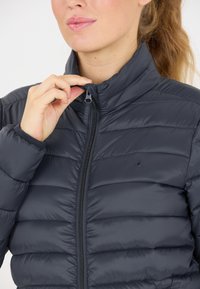 Dunkelgraue gepolsterte Jacke mit Stehkragen und Reißverschluss. Merkmale sind horizontale Nähte und eine weiche, glatte Textur. Keine sichtbaren Logos.