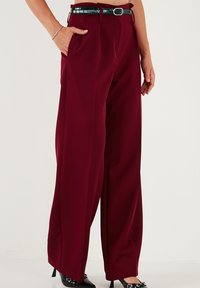 Pantaloni largi burgundy cu o textură fină, prevăzuți cu buzunare laterale, talie înaltă și o curea lucioasă neagră. Formă lungă și fluidă.